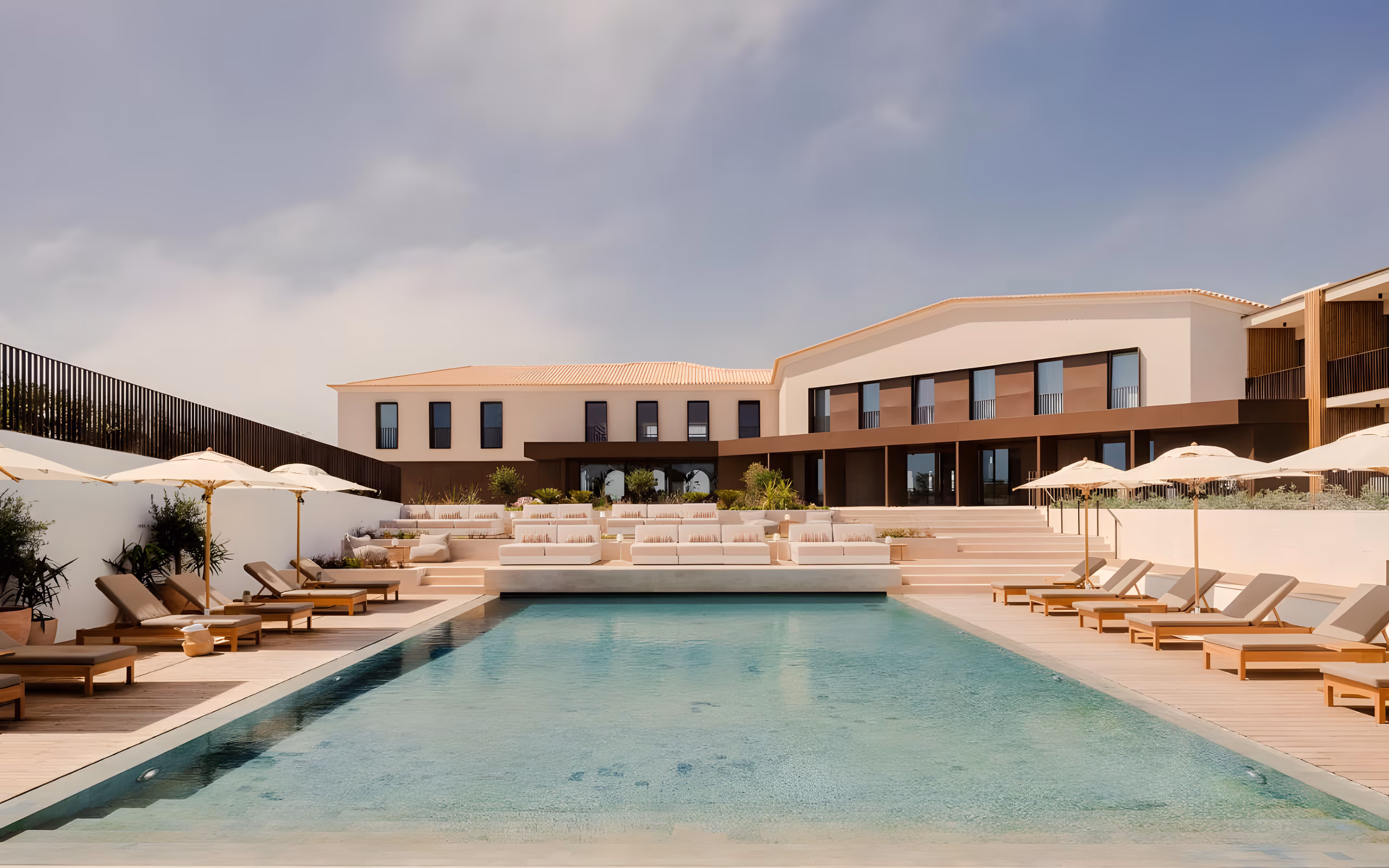 Long outdoor pool lined with loungers and parasols below a low white clifftop building