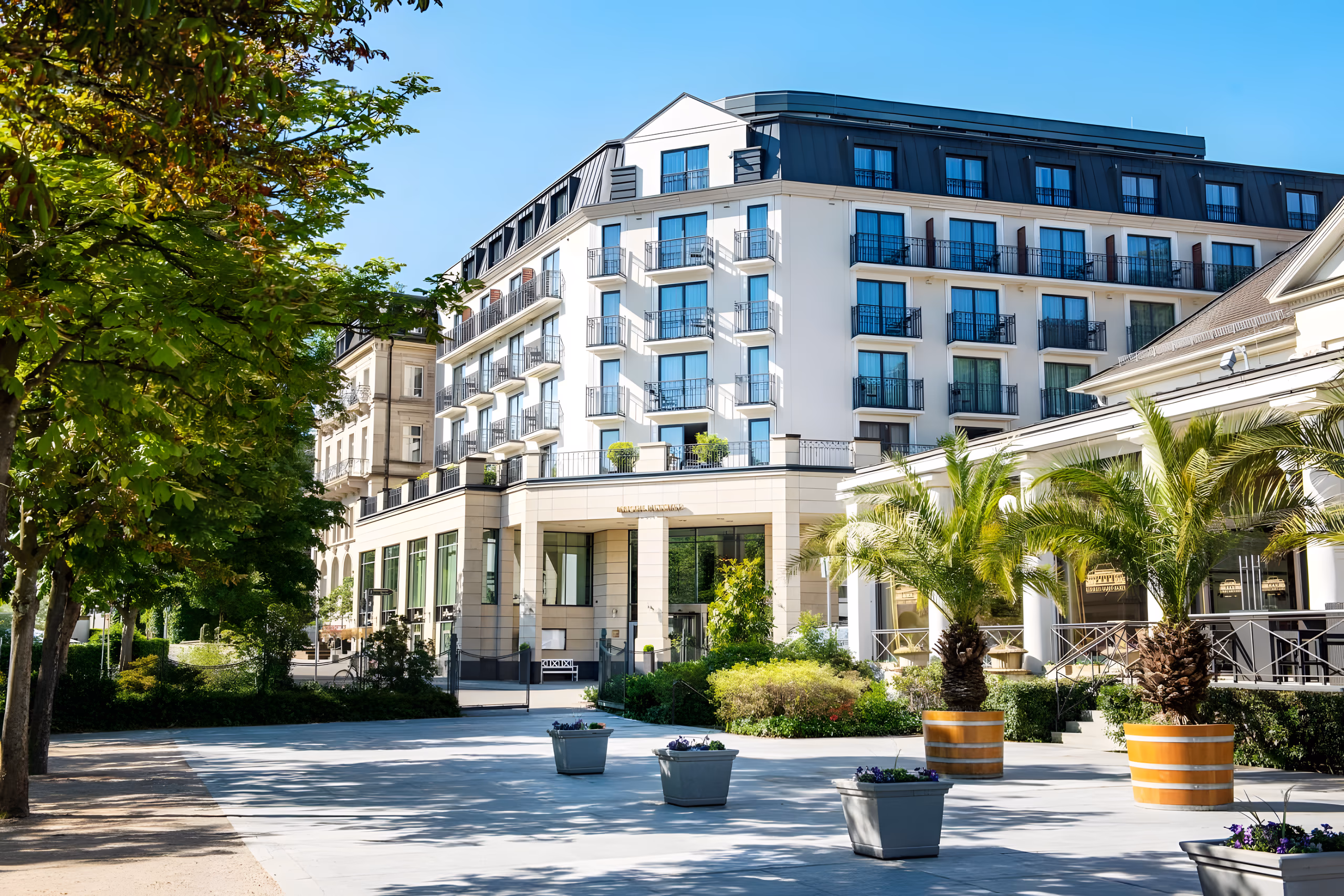 Belle Epoque facade with balconies and palm-lined forecourt on a tree-shaded spa town promenade